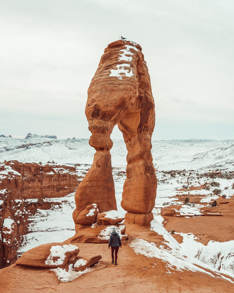 female hiker walking under delicate arch in arches national park utah in winter