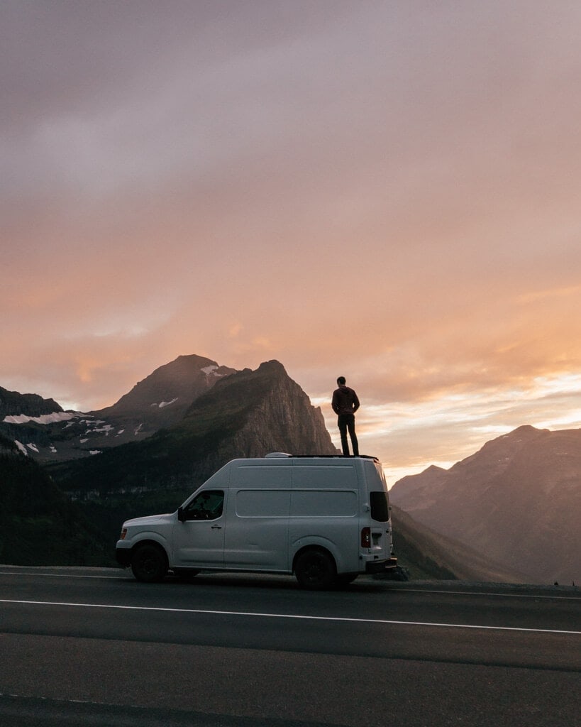 male standing on top of a white campervan while watching a vibrant pink sunset over going to the sun road glacier np